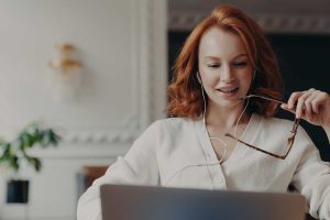 young woman with red hair, holding glasses in her left hand looks at her laptop computer.